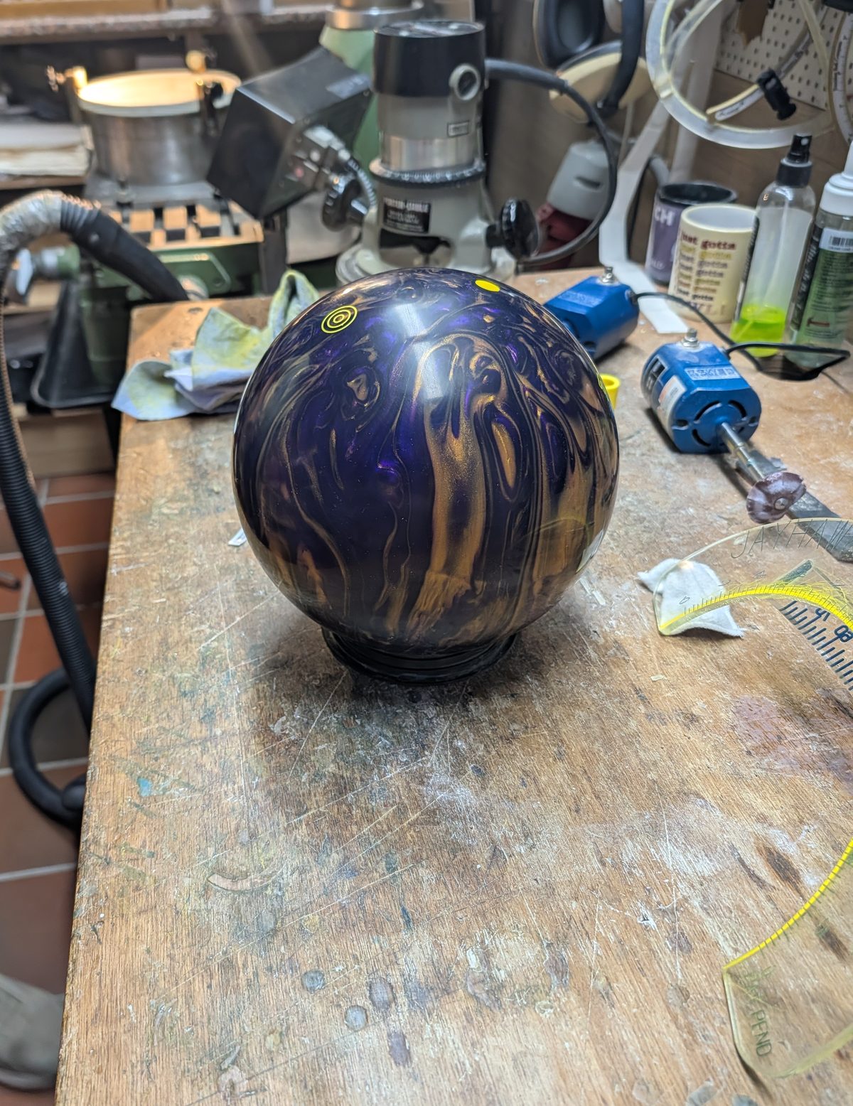 Freshly drilled purple and gold Ebonite Fireball bowling ball on a pro shop workbench next to the drill press and a measuring tape